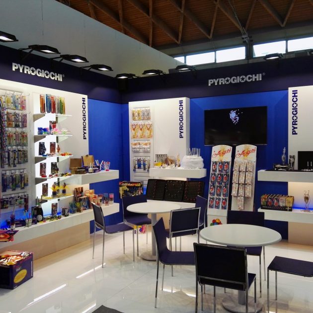 Stand espositivo Pyrogiochi con prodotti colorati su scaffali e tavoli con sedie, ambiente illuminato.