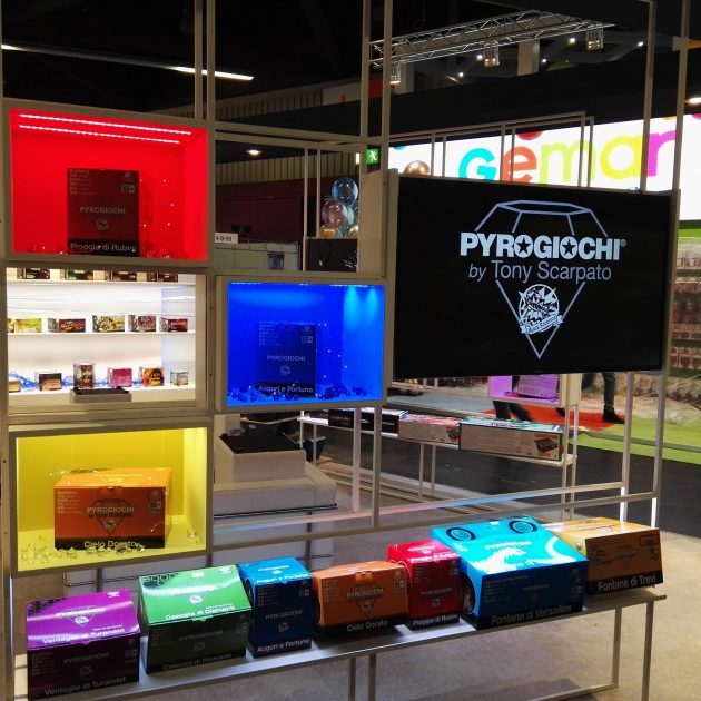 Stand fieristico con giochi pirotecnici colorati esposti su scaffali illuminati al chiuso.