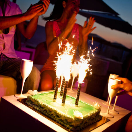 Festa serale con torta di compleanno, scintille e diverse persone che applaudono, bicchieri in primo piano.