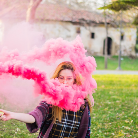 Una donna con occhiali crea un effetto visivo con fumo rosa in un parco.