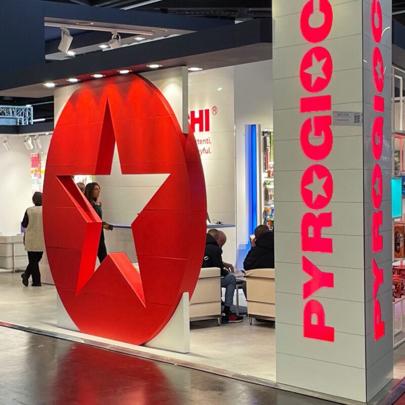 Stand fieristico con grande stella rossa, persone che camminano, espositori illuminati e scritta PYROGIOCHI.