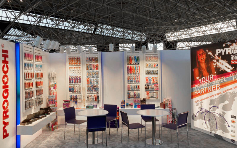 Stand espositivo Pyrogiochi, con tavoli, sedie e scaffali di prodotti colorati sotto un tetto di metallo e vetro.