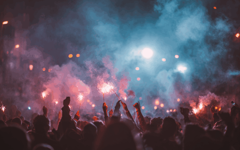 Folla di persone durante una festa notturna con tante luci e fumi colorati in atmosfera festosa.