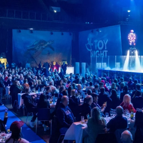 Header_Rahmenprogramm Una folla seduta a tavoli guarda uno spettacolo intitolato Waterworld in un grande auditorium.