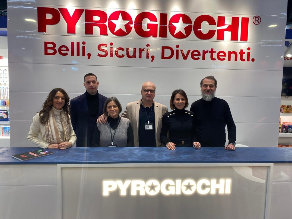 Sei persone sorridenti posano davanti a uno stand Pyrogiochi con il motto Belli, Sicuri, Divertenti.