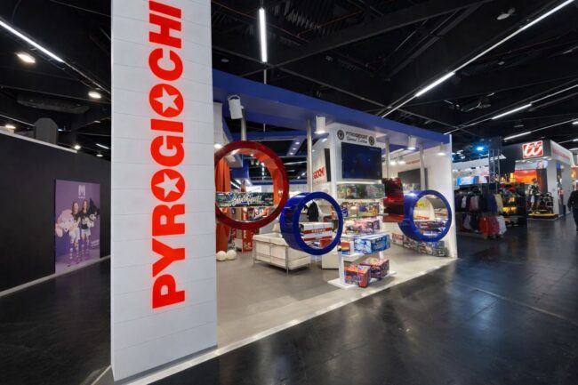 Negozio PyroGiochi in fiera, espositori colorati e giochi in mostra sotto un'illuminazione intensa.